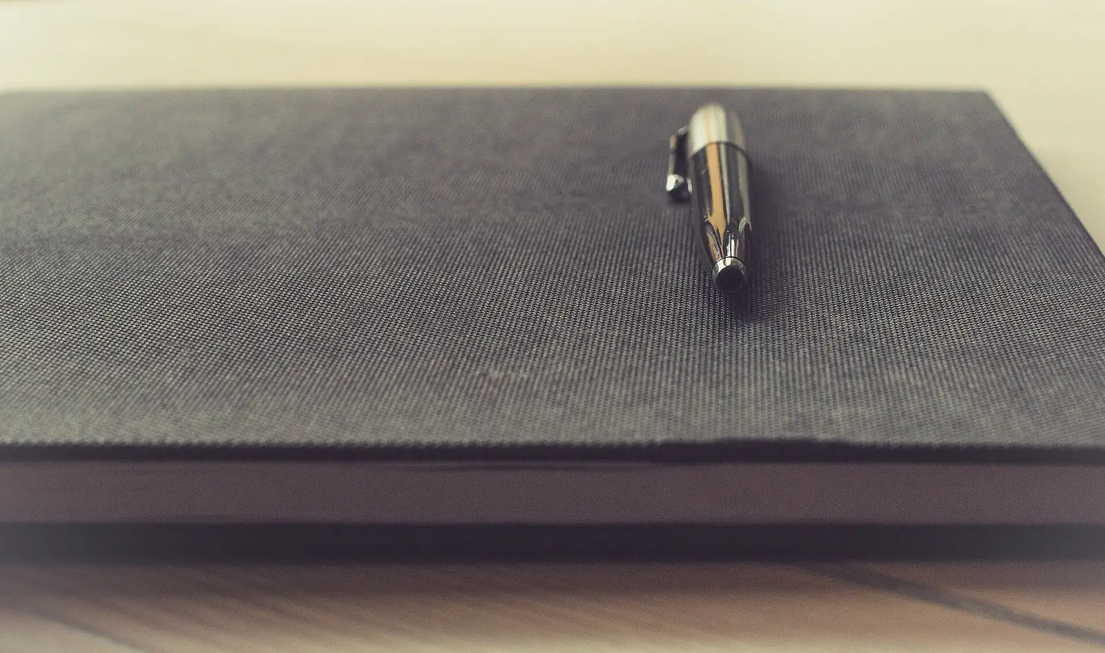A silver pen resting on a closed dark grey notebook on a wooden desk